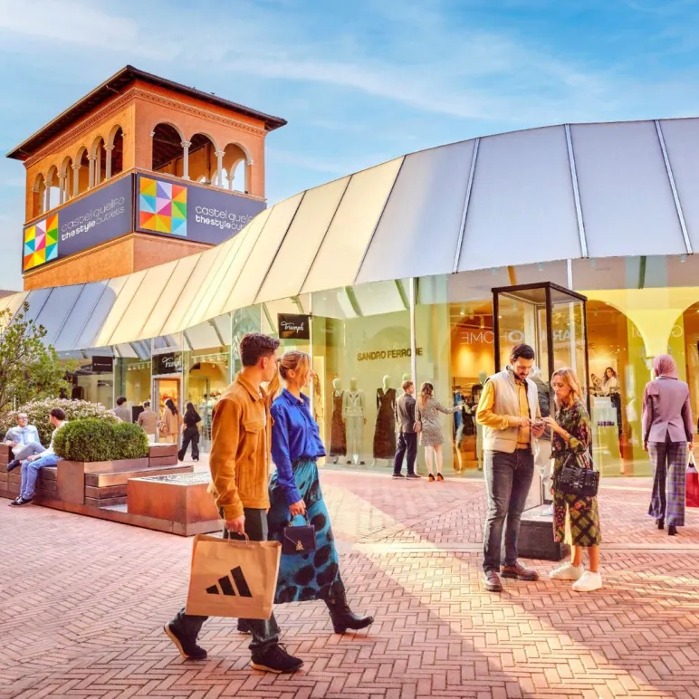 Persone che passeggiano e fanno shopping all’aperto a Castel Guelfo The Style Outlets, con le vetrine dei negozi sullo sfondo e la torre con il logo del centro